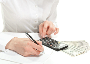 A person using a calculator and writing on paper with several U.S. dollar bills placed beside them on a white surface.