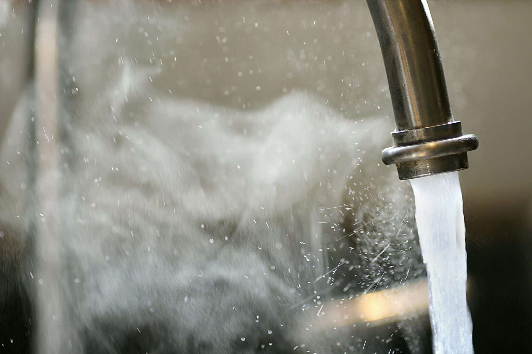 Hot steaming running tap water is pouring out of a stainless steel kitchen faucet.