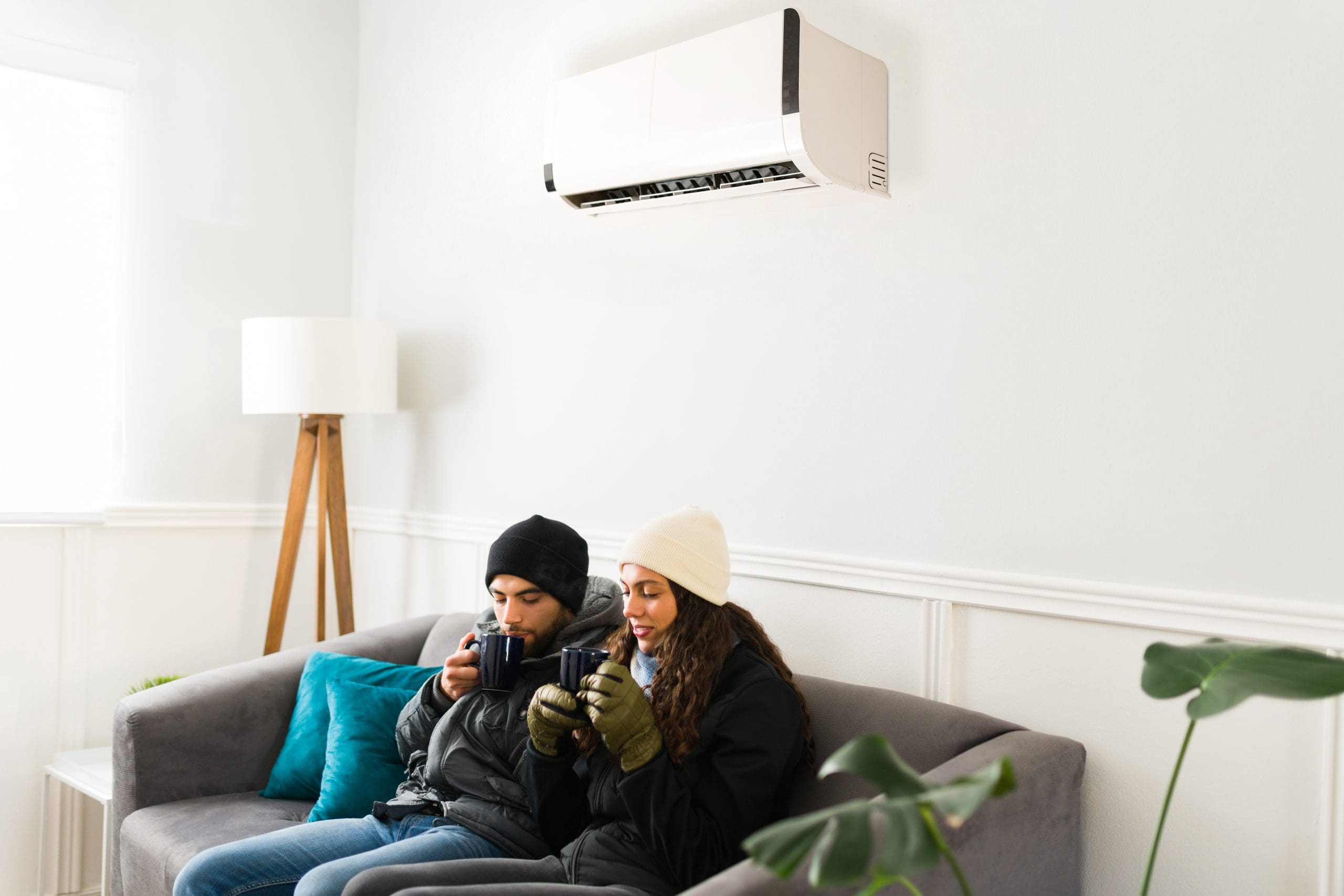 Young couple beneath air conditioner in home with warm clothes on bundled up