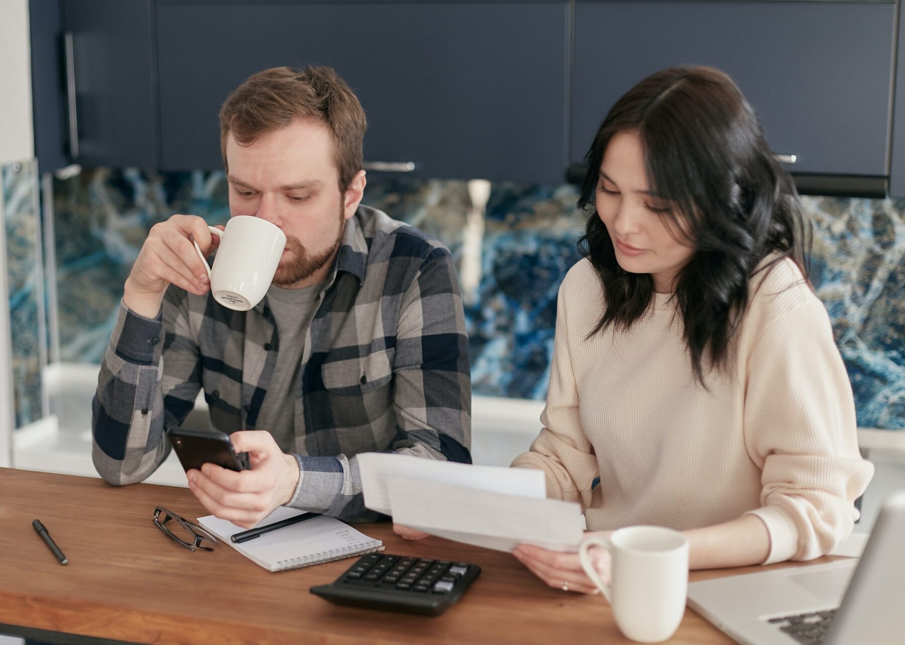 Husband and wife sit at table going over bills while husband drinks a cup of coffee.