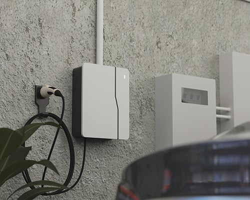 A wall-mounted electric vehicle (EV) charger installed in a garage or outdoor area. A black charging cable is connected, and part of a car is visible in the foreground. The setup appears clean and modern.
