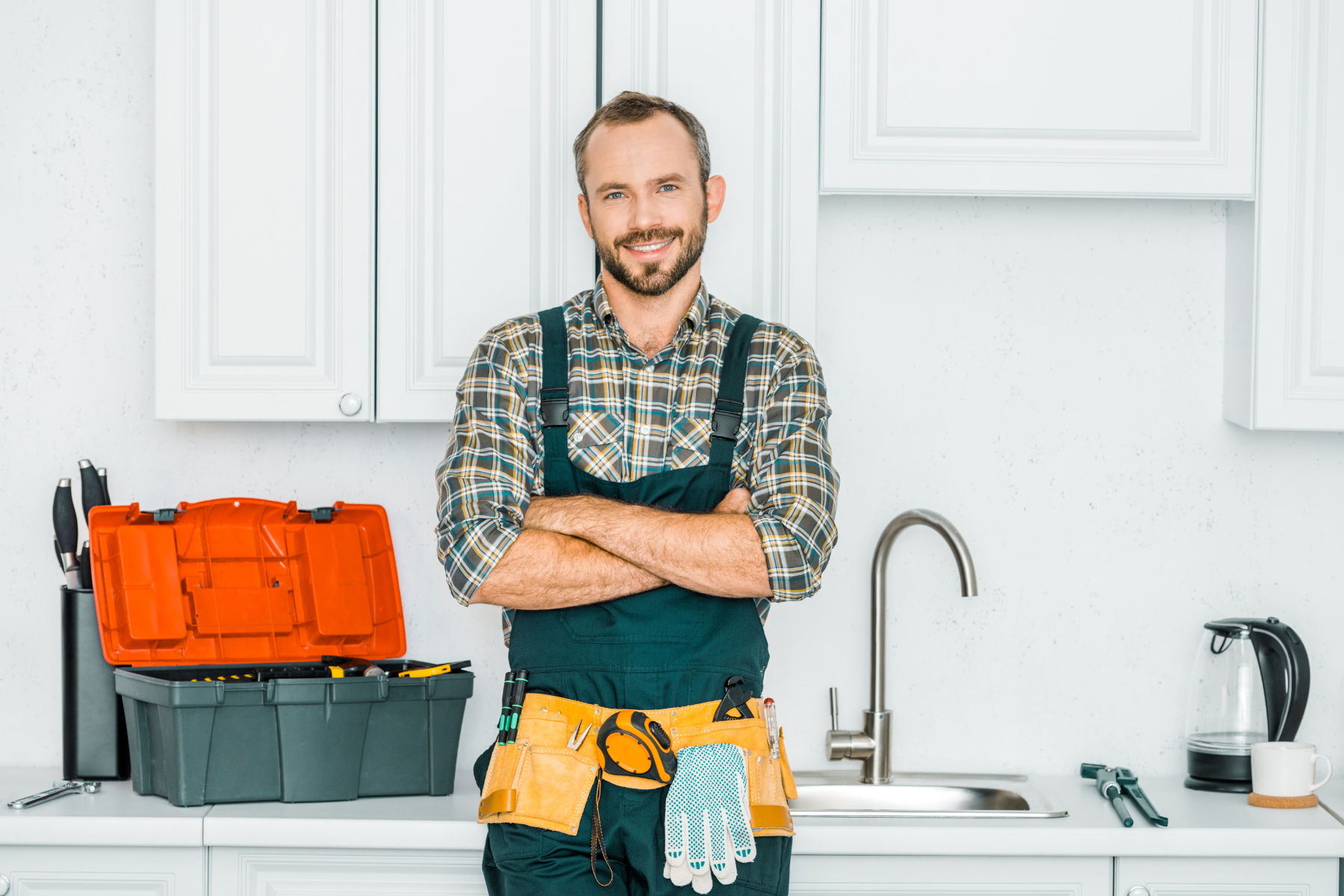 A smiling plumber wearing a tool belt and work overalls stands in a bright kitchen with arms crossed. Behind him is an open toolbox with tools, a modern sink, and a kettle on the countertop