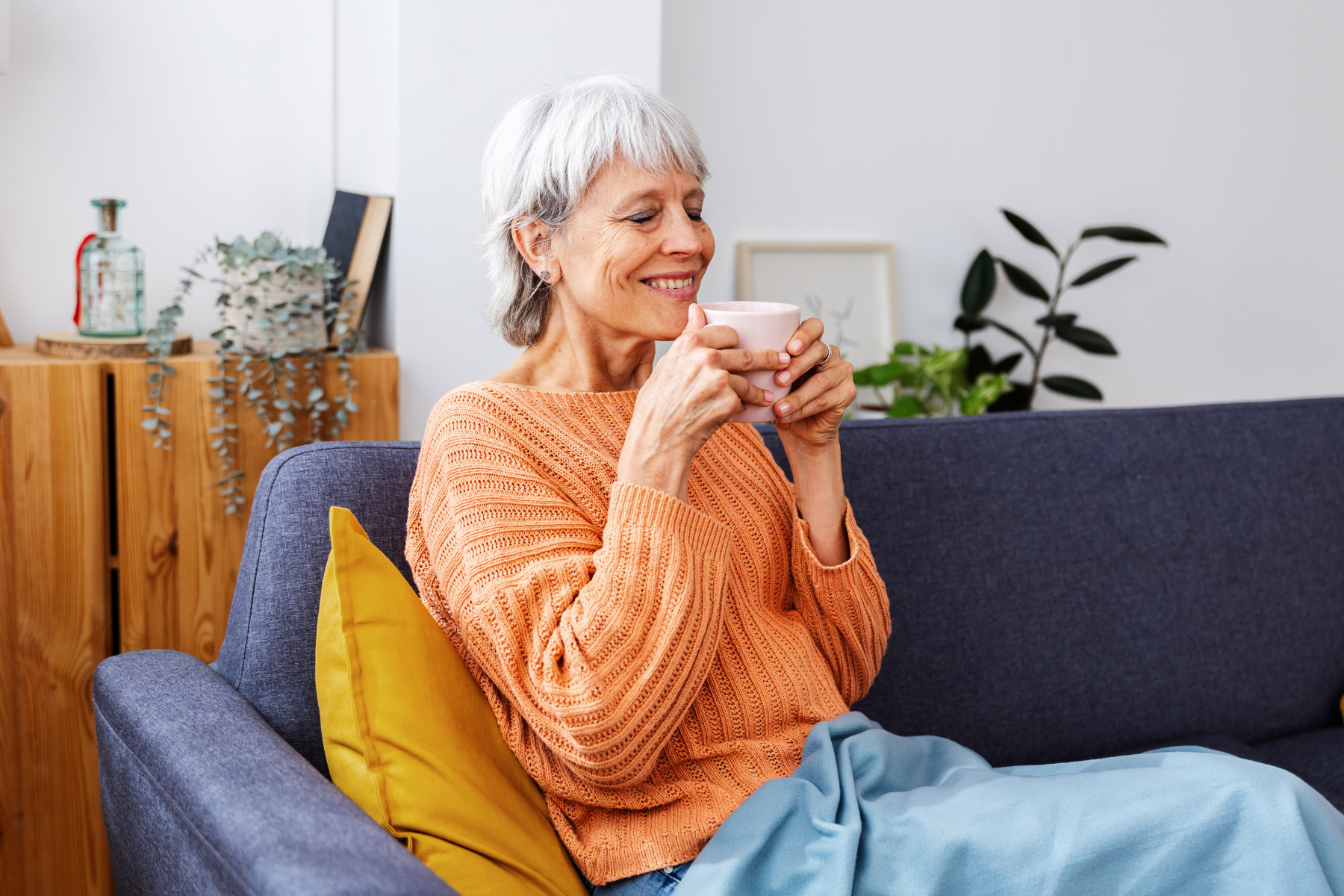 An older woman sitting on a sofa, smiling contentedly while holding a warm cup of tea. She is wrapped in a light blue blanket, wearing an orange sweater, with indoor plants and wooden decor in the background, enjoying the warmth of her home.
