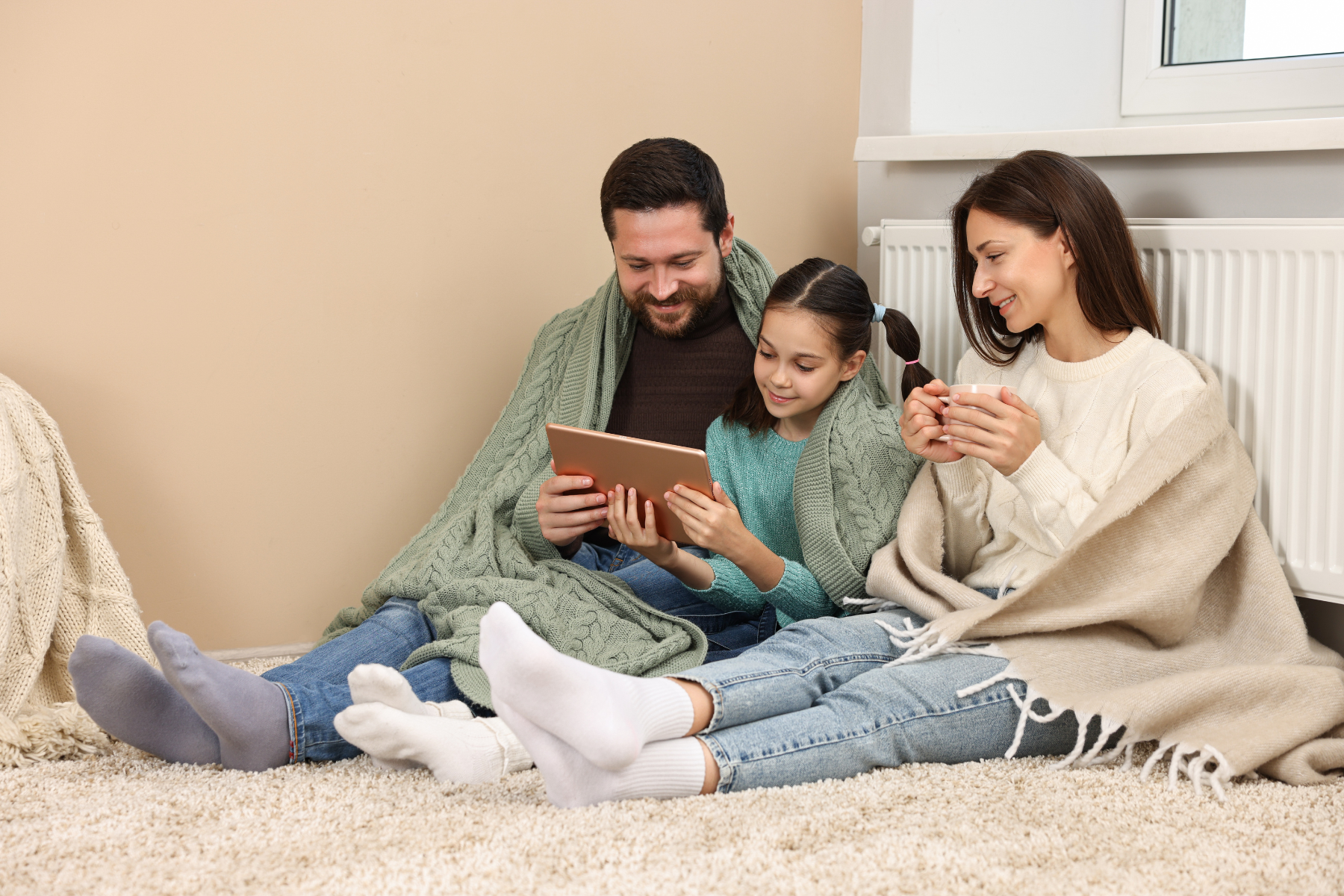 A smiling family of three, wrapped in blankets, sitting on a carpeted floor in front of a radiator. The father and daughter are looking at a tablet while the mother holds a warm drink, enjoying a cozy moment despite a furnace failure.
