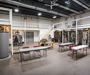 An industrial-style workspace with high ceilings, featuring tables and chairs arranged in the center and various equipment and machinery along the walls. A staircase leads to a mezzanine level with railings, and the area is well-lit with overhead lights.