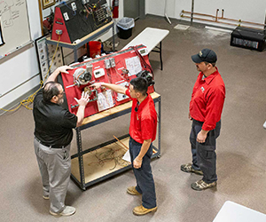 Three people gathered around a red training board equipped with wiring and electrical components in a workshop or classroom setting. Two of them wear red shirts, and one wears a black shirt, as they discuss or examine the setup.