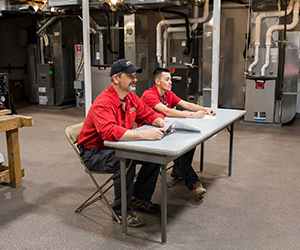 Two people wearing red shirts sitting at a table in an industrial training room, surrounded by HVAC equipment and metal ductwork.