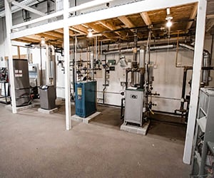 Several large HVAC and plumbing systems installed along a wall in an industrial training area, with exposed pipes, valves, and equipment beneath a wooden mezzanine structure.
