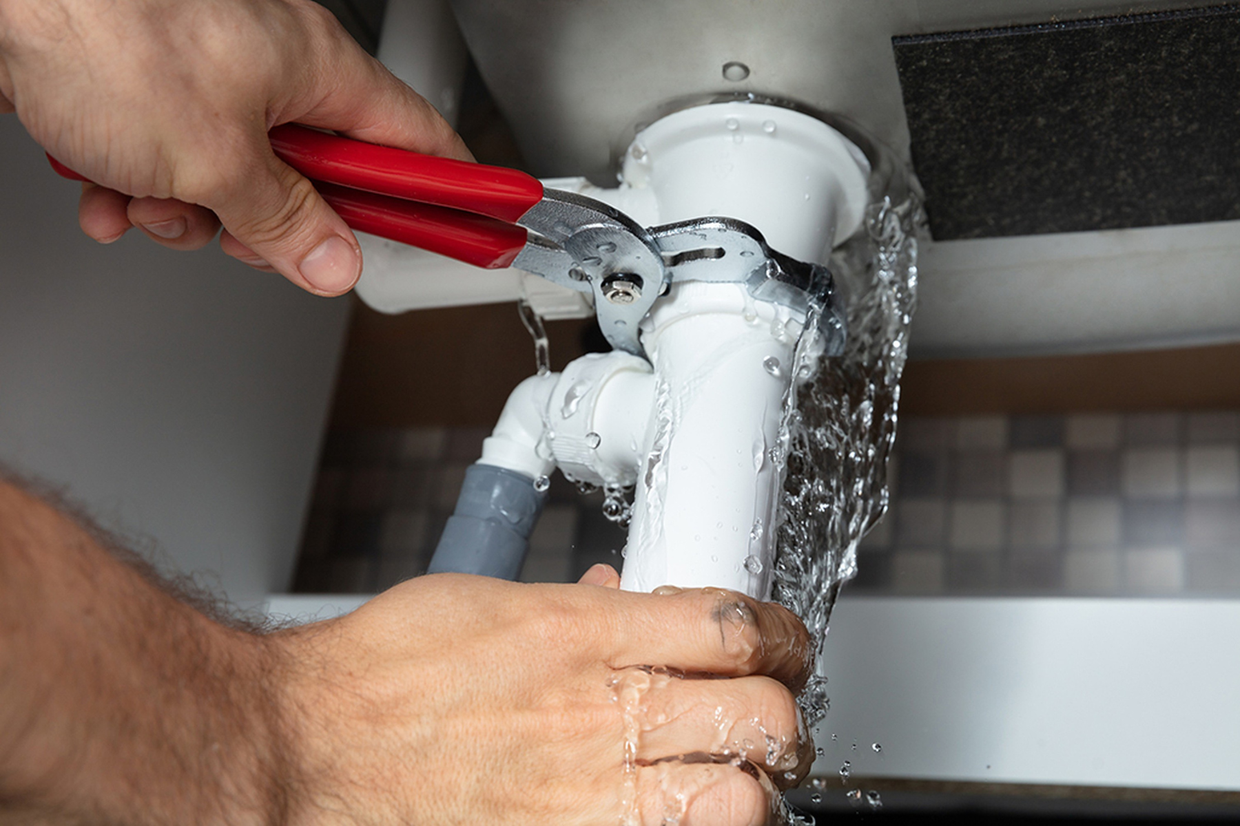 A close-up of a plumber's hands using a wrench to fix a leaking pipe under a sink, with water spilling out.