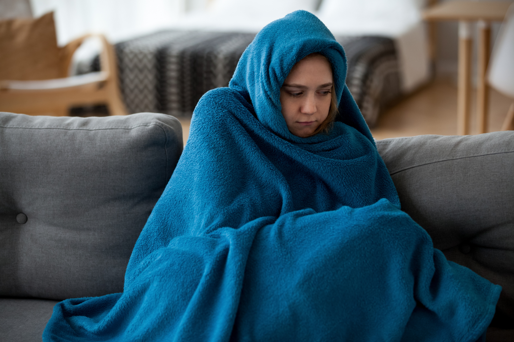 woman sitting on a gray sofa, wrapped in a thick blue blanket, with a somber expression. The room appears cozy, with soft lighting and blurred furniture in the background.