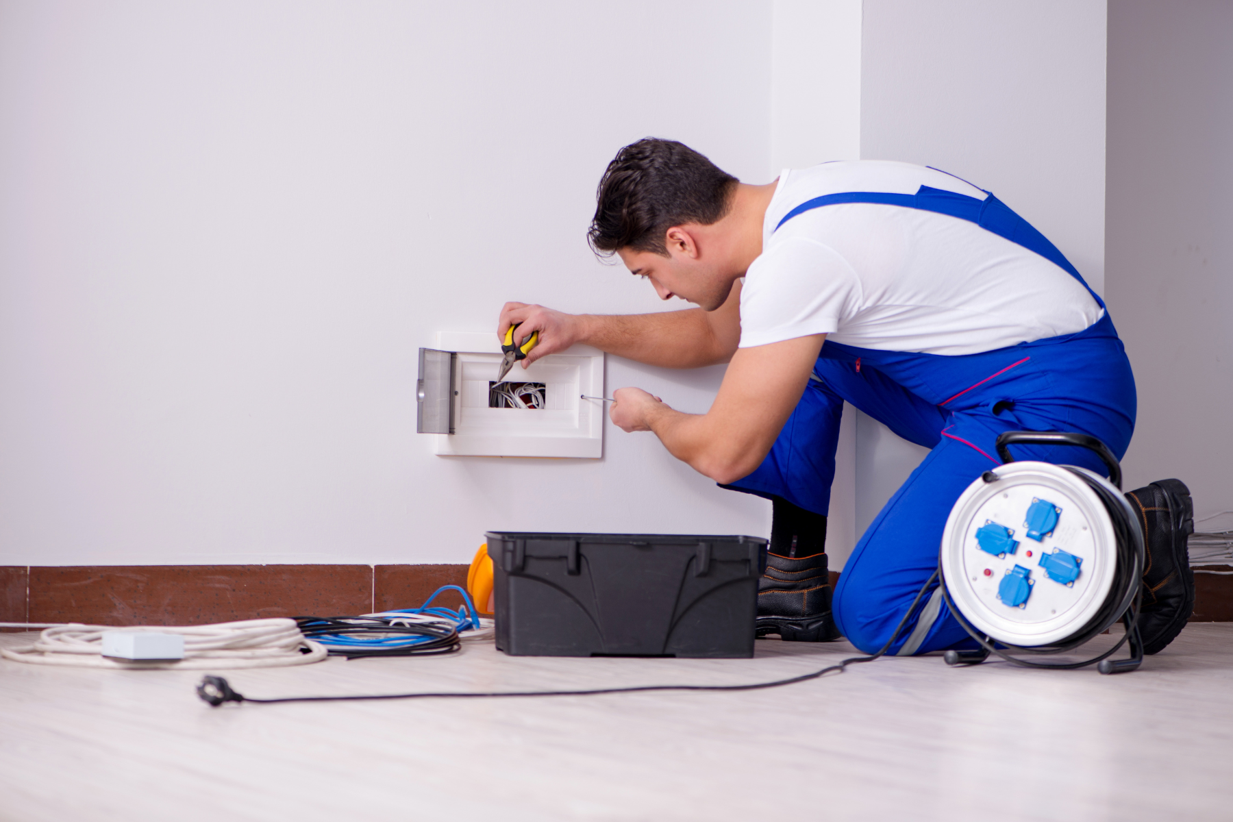 A man wearing a blue overall, working on an electrical panel installed on a wall. Tools, cables, and an electrical cord reel are visible around him.