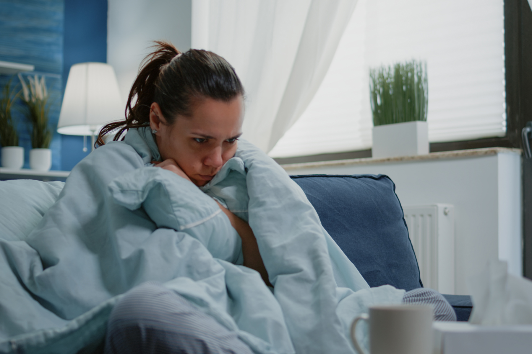 A woman sitting on a blue sofa wrapped in a light blue blanket, with a distressed expression, and a window with white blinds and indoor plants in the background.
