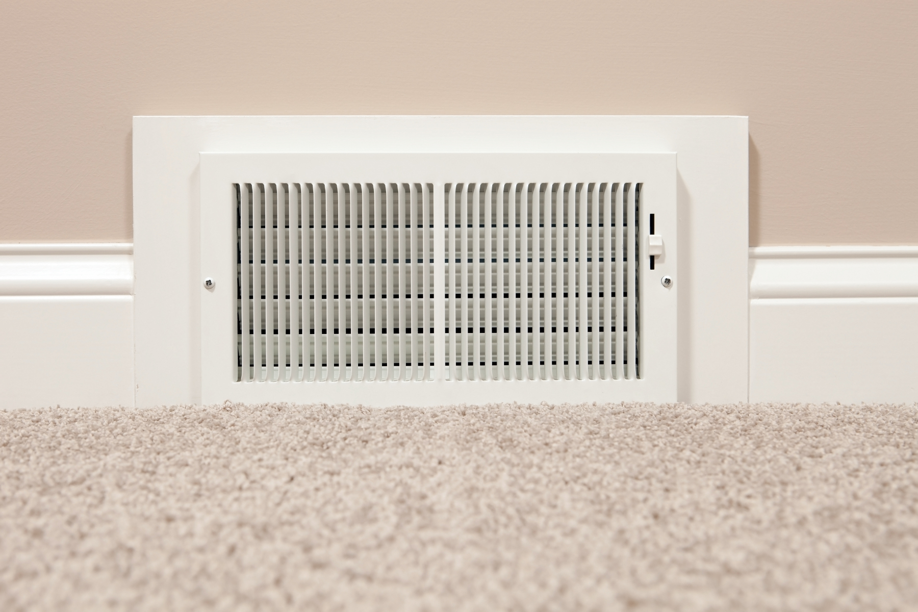 A white rectangular air vent is mounted at the base of a beige wall, just above a soft, beige carpet. The vent has vertical slats and a small lever on the right side for adjusting airflow.