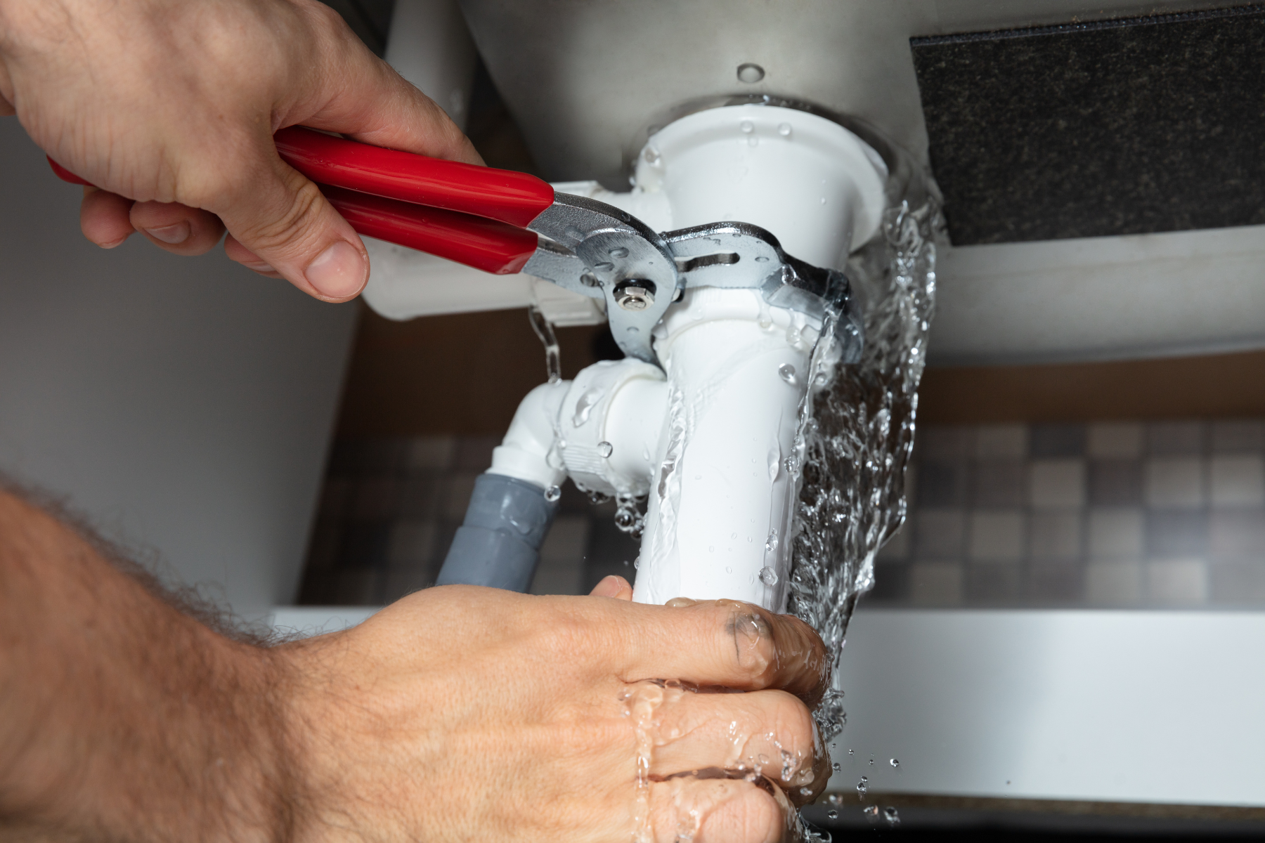 A person is holding a pair of red-handled pliers and attempting to fix a leaking white plastic pipe under a sink. Water is visibly spraying out from the joint, and the surrounding area includes other pipes and fittings