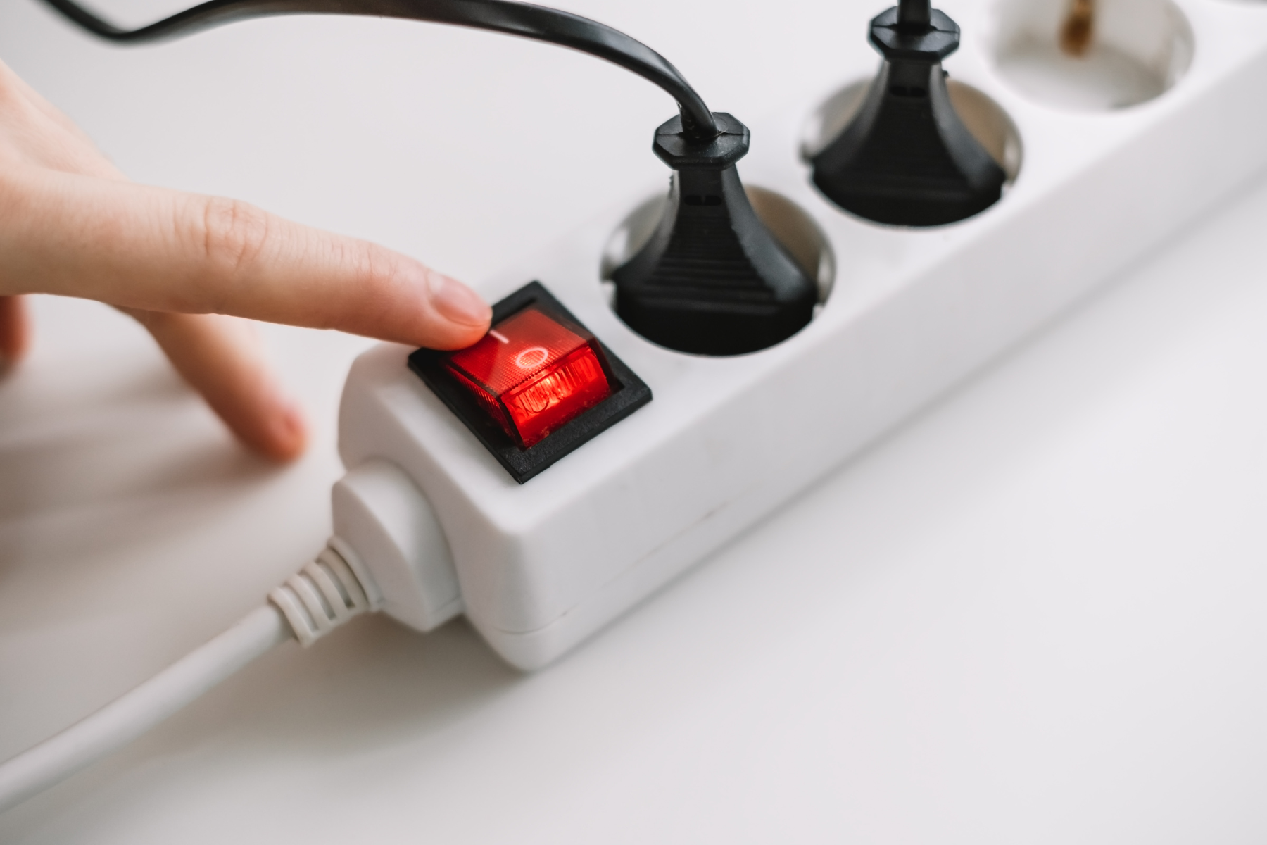 A close-up of a hand reaching to press the illuminated red switch on a white power strip. Two black plugs are inserted into the strip, and the scene is set against a plain white surface.