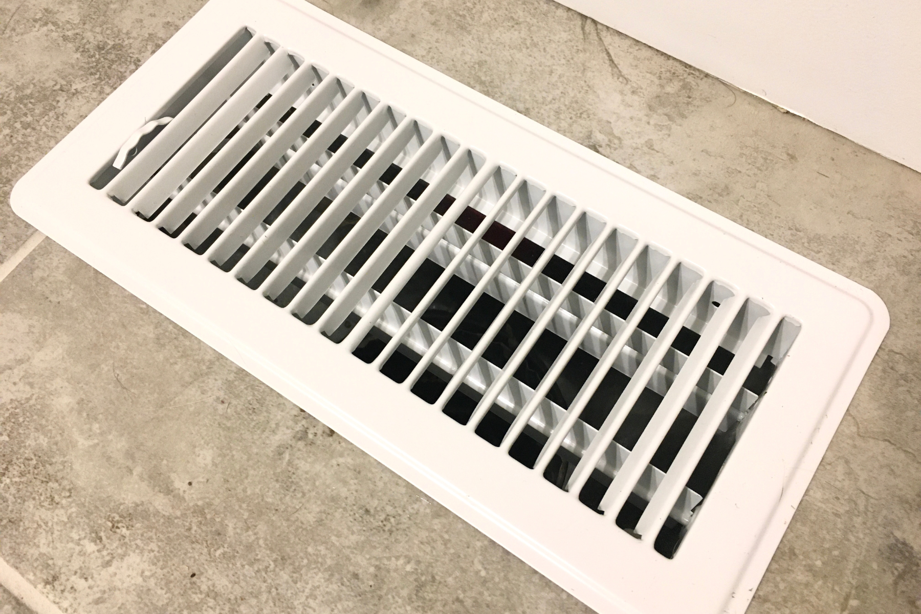 A white rectangular floor vent is installed on a tiled surface. The vent has angled slats and appears clean and flush with the floor. Part of a white wall is visible behind the vent.