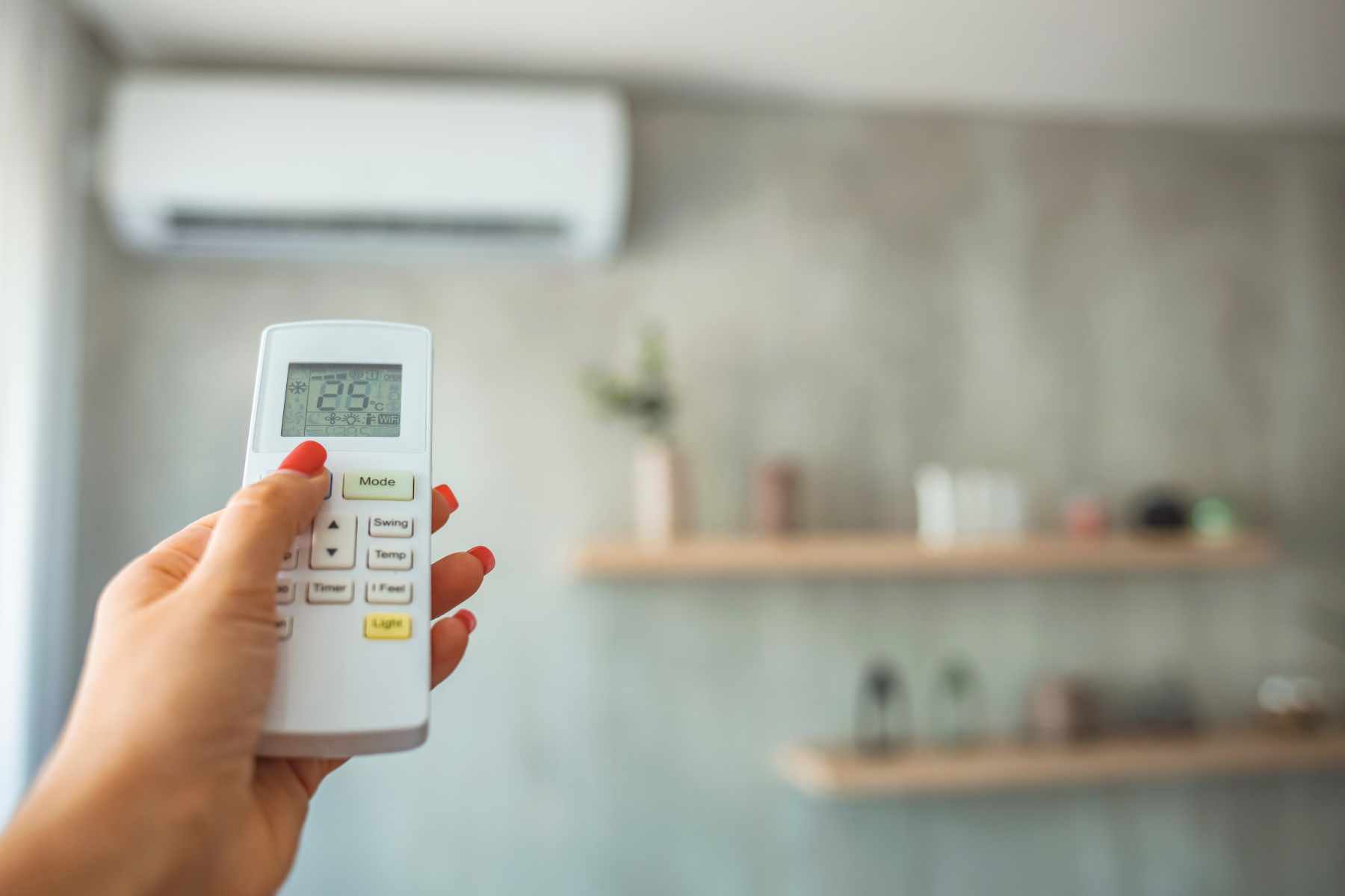 A hand holding a remote control for an air conditioning unit. The display on the remote shows the current temperature as 26°C, with buttons for adjusting modes, temperature, and fan settings.