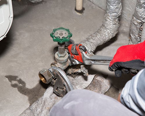 Hands wearing red and black gloves using two wrenches to tighten or loosen a pipe connection near a green-handled valve, with insulated pipes and a concrete floor in the background.