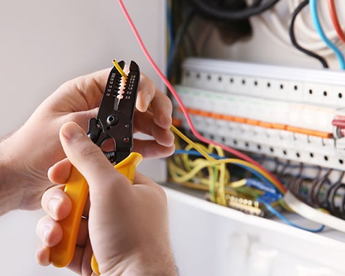 A person carefully strips the insulation from an electrical wire using yellow-handled wire strippers. Multiple colored wires are visible inside an electrical panel, indicating work related to wiring or electrical installation.