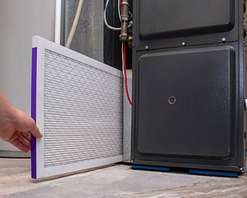 A hand placing or removing a rectangular air filter beside a large black furnace unit in a utility or basement area.