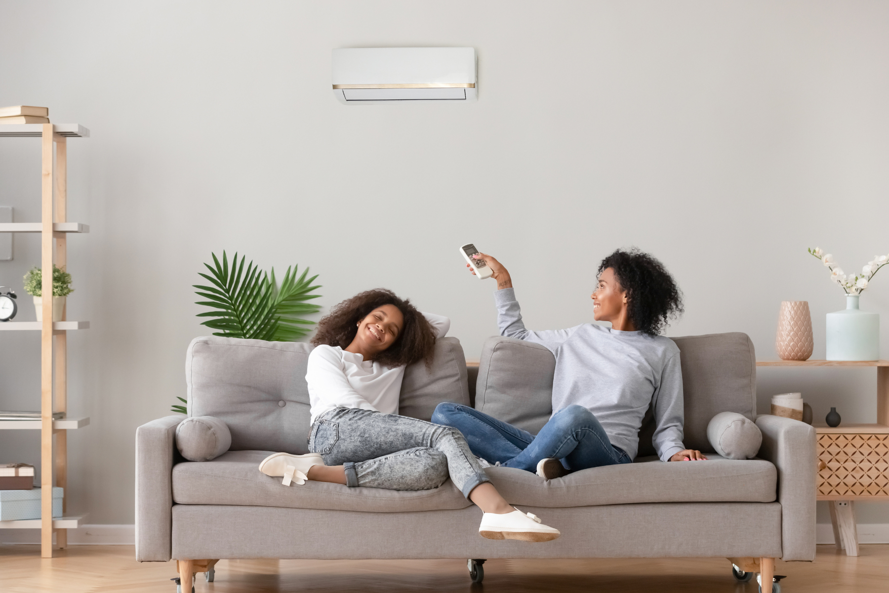 Two women relax on a couch under a wall-mounted AC, enjoying cool air indoors.