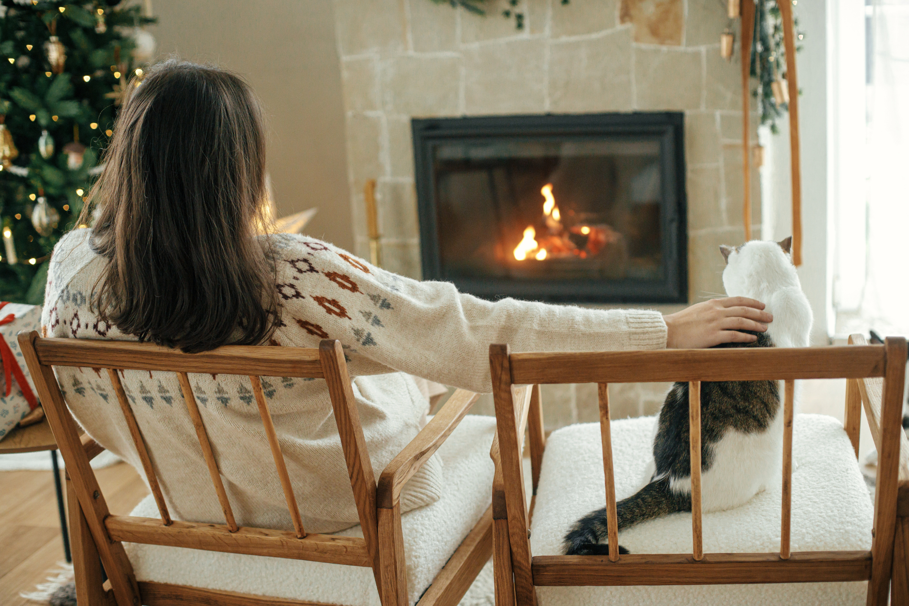 A woman in a cozy sweater sits in a wooden chair, gently petting a cat while enjoying the warmth of a lit fireplace.