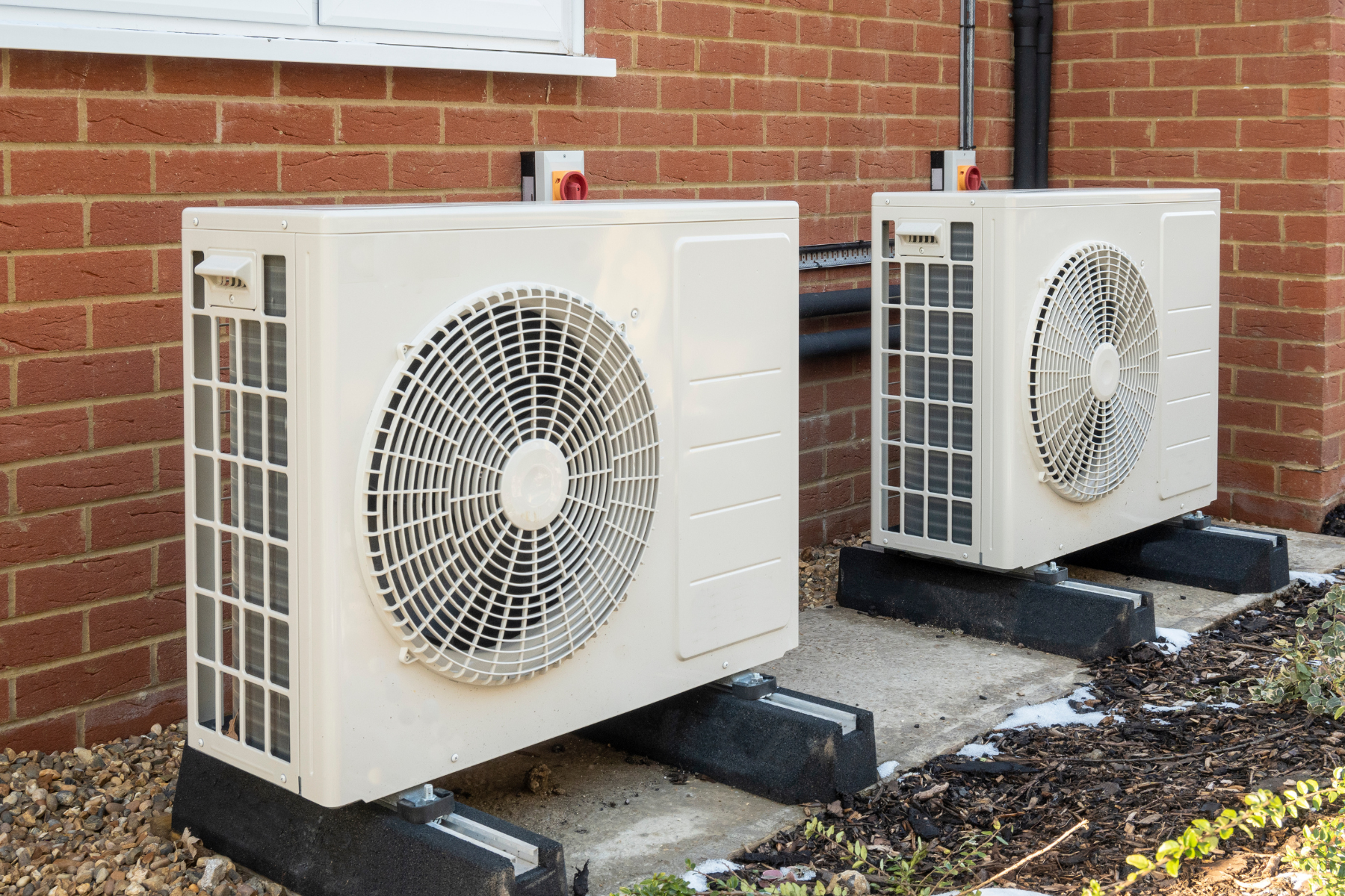 Two outdoor air conditioning units are installed on a concrete base against a brick wall.