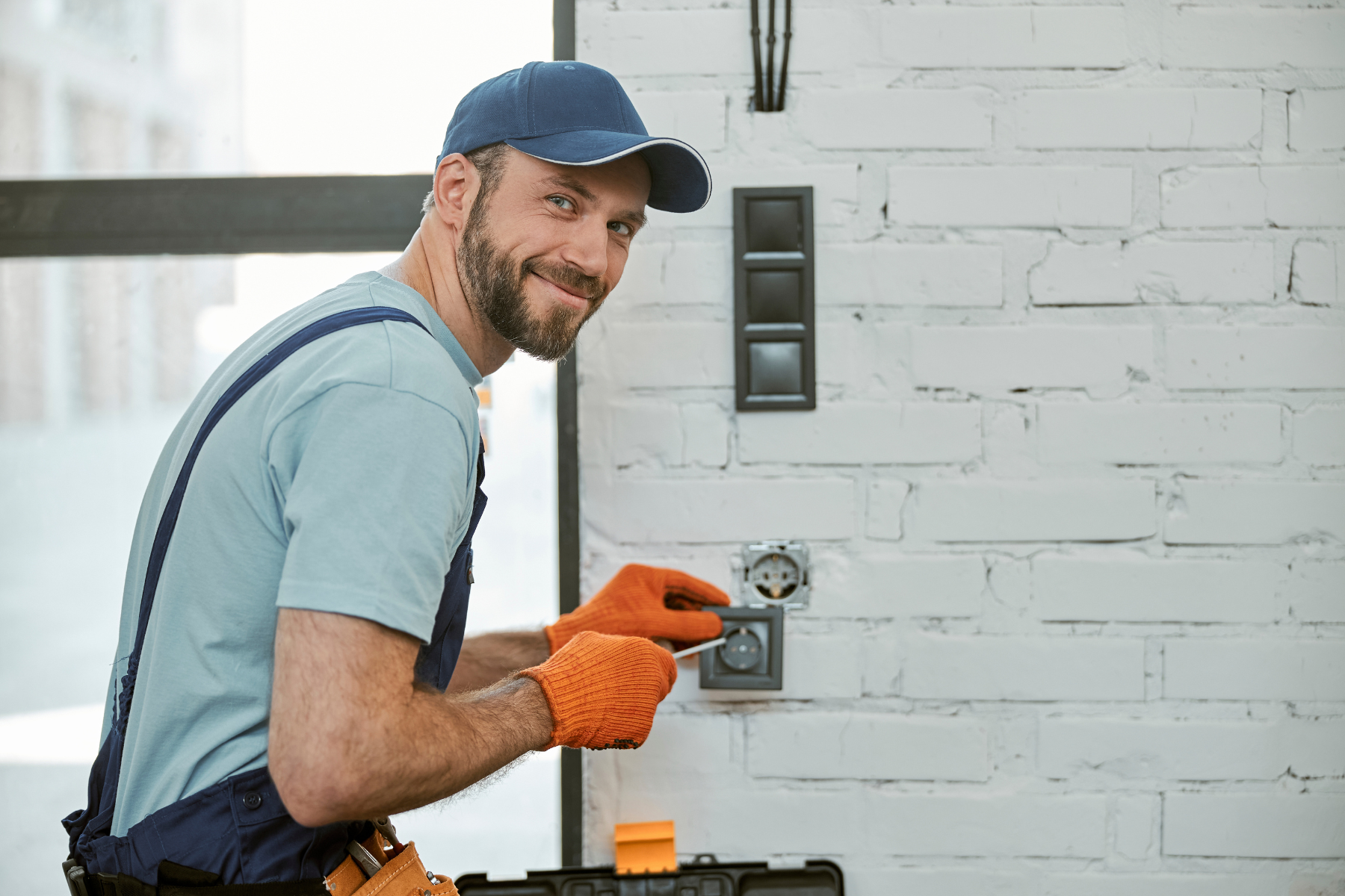 An electrician installs or repairs a wall outlet, using a screwdriver while smiling at the camera.