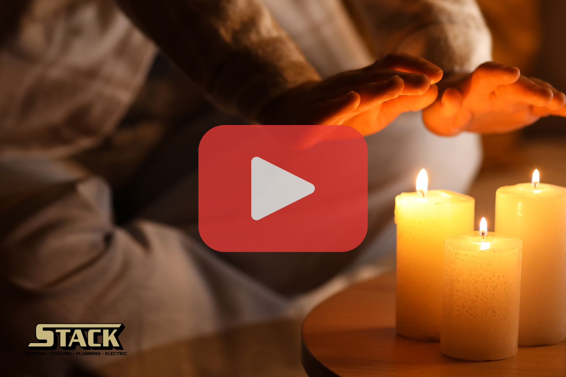 A person sits indoors with their hands held out toward three lit candles on a small table, using the flames for warmth. The scene conveys a lack of central heating or a heating failure, suggesting the person is trying to stay warm using candles as a temporary heat source. The setting appears dim and cozy but also highlights vulnerability to cold temperatures.