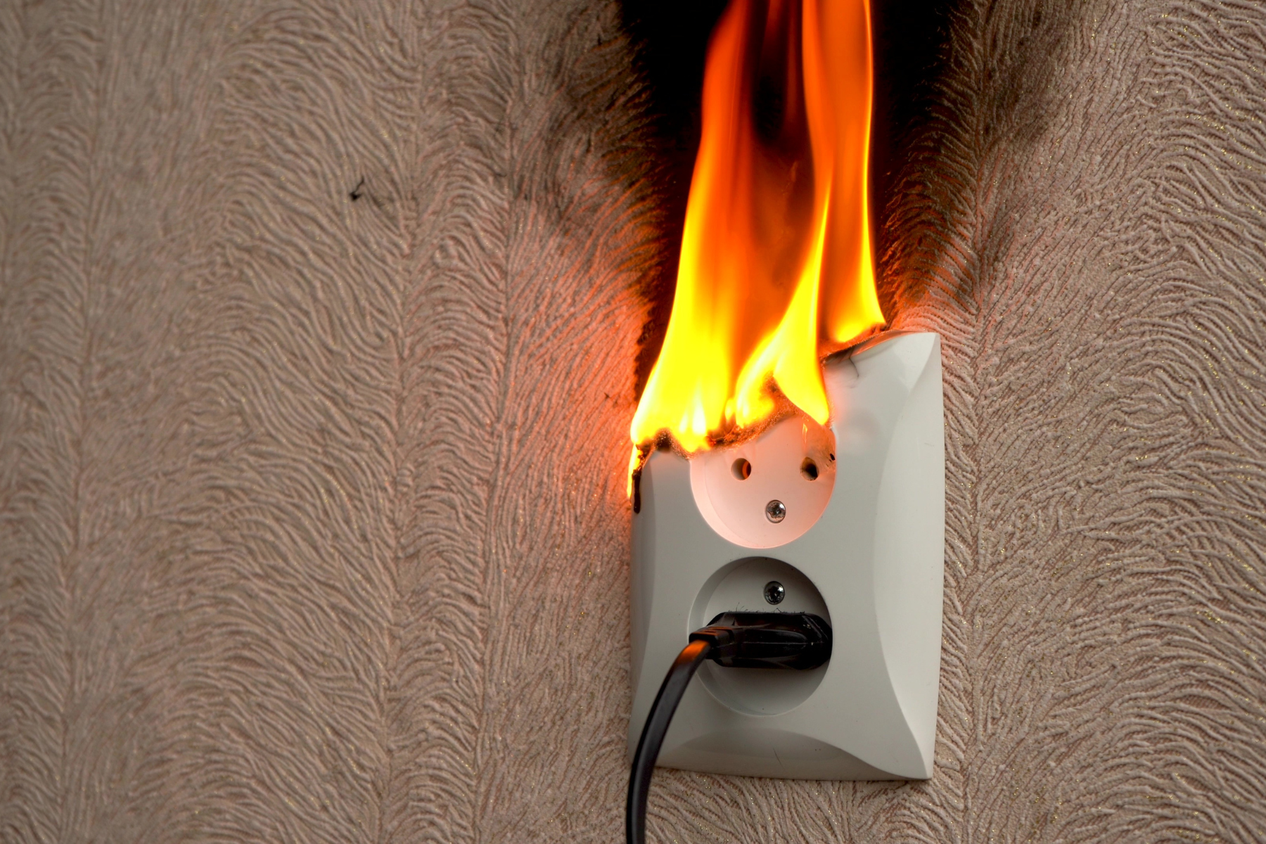 Flames are rising from an electrical wall outlet where a plug is inserted. The heat has scorched the plastic outlet cover and is actively burning, indicating a serious electrical fire hazard.