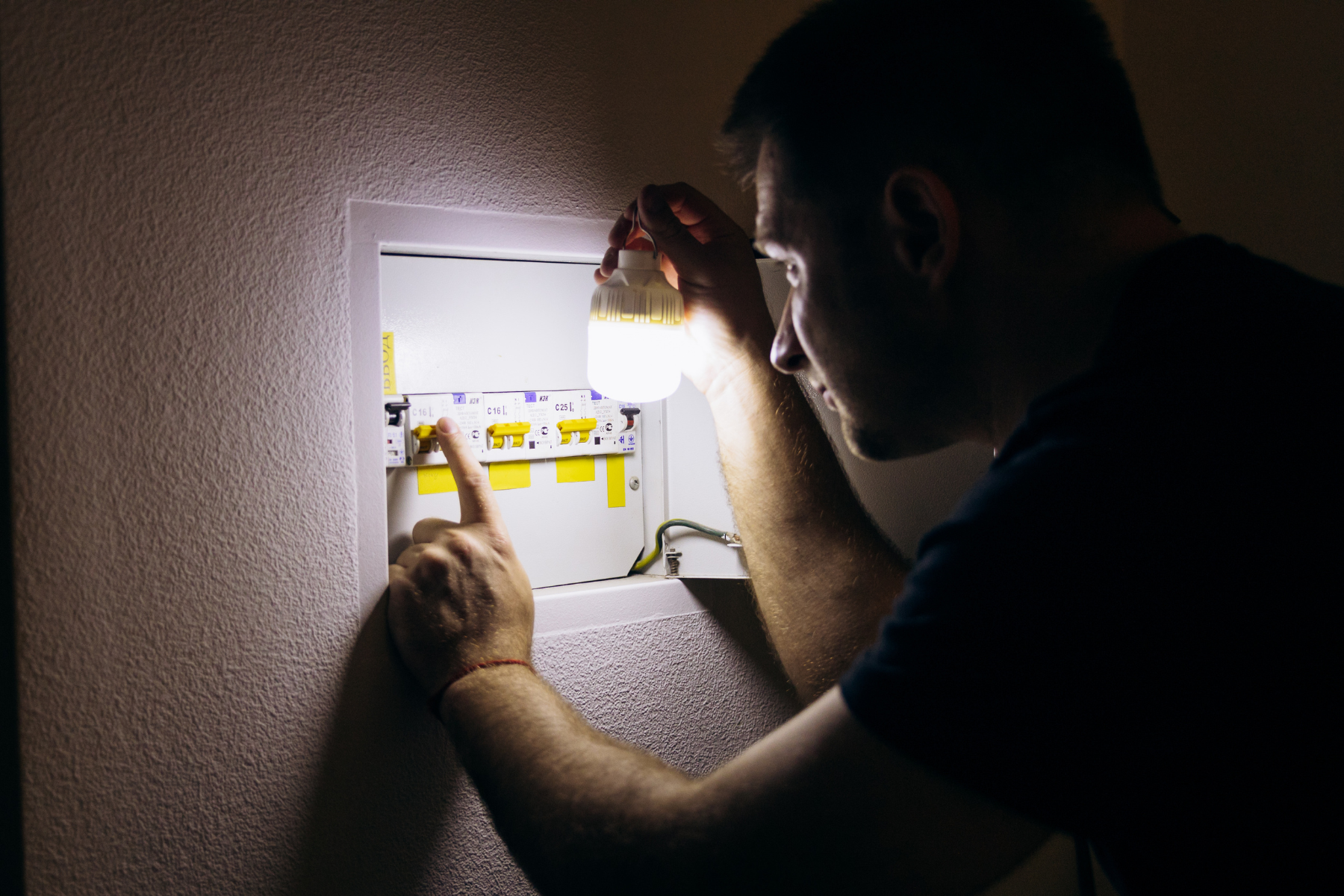 A person shines a portable light into an open electrical breaker panel while pointing at a specific switch.