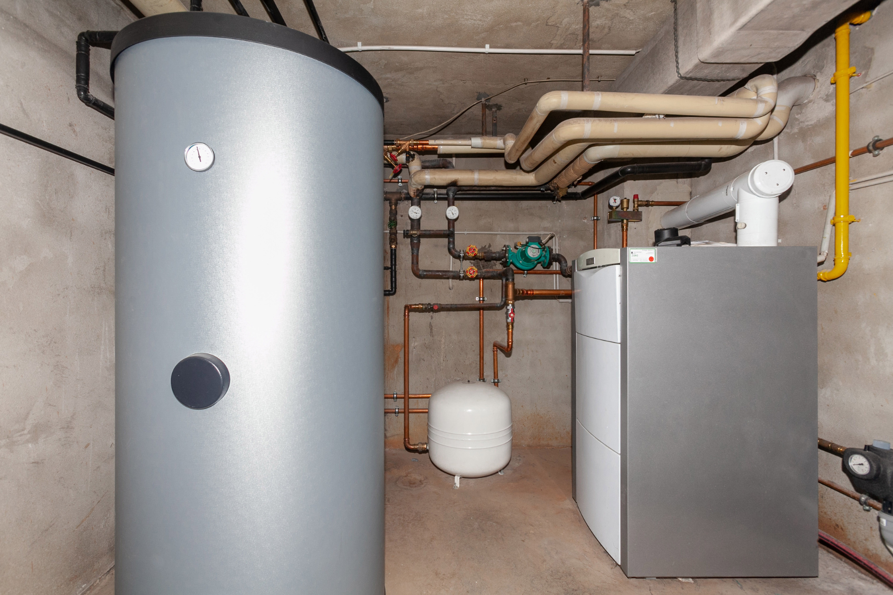 A large cylindrical hot water storage tank and a modern boiler connected by insulated pipes, valves, and gauges in a concrete utility room.