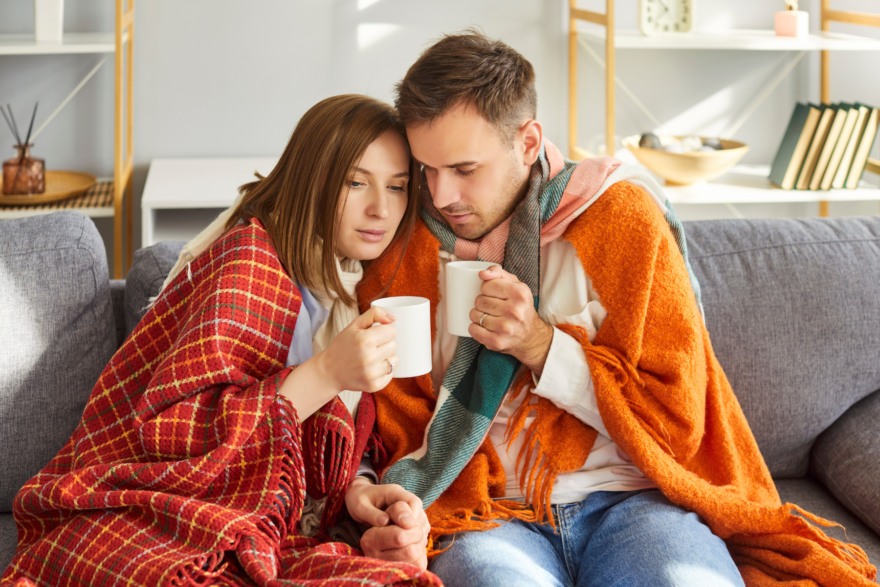 A couple sits closely wrapped in cozy blankets and holding warm drinks, suggesting they are trying to stay warm in a chilly home.