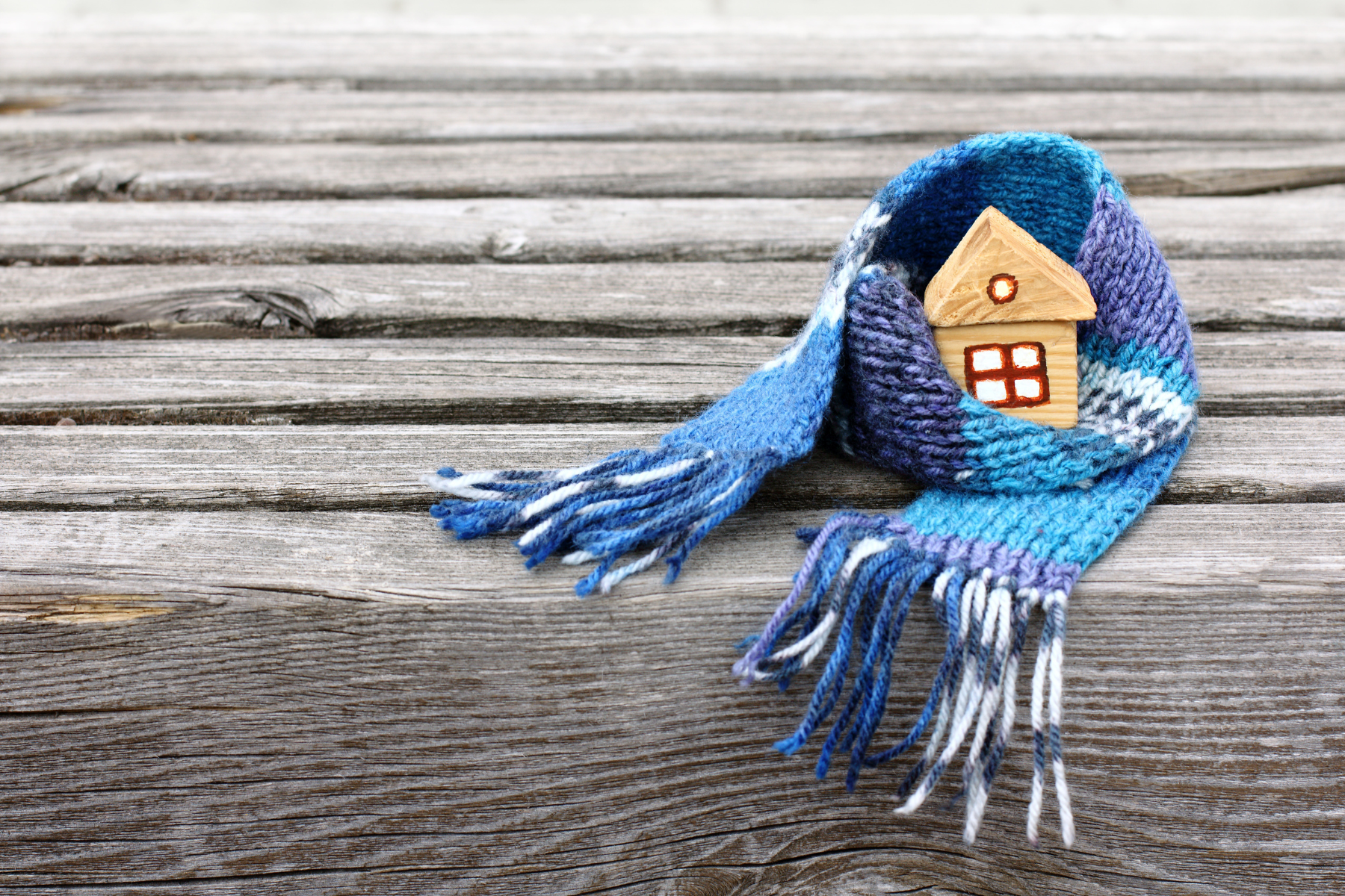 A tiny wooden house wrapped snugly in a blue knitted scarf rests on a rustic wooden surface.