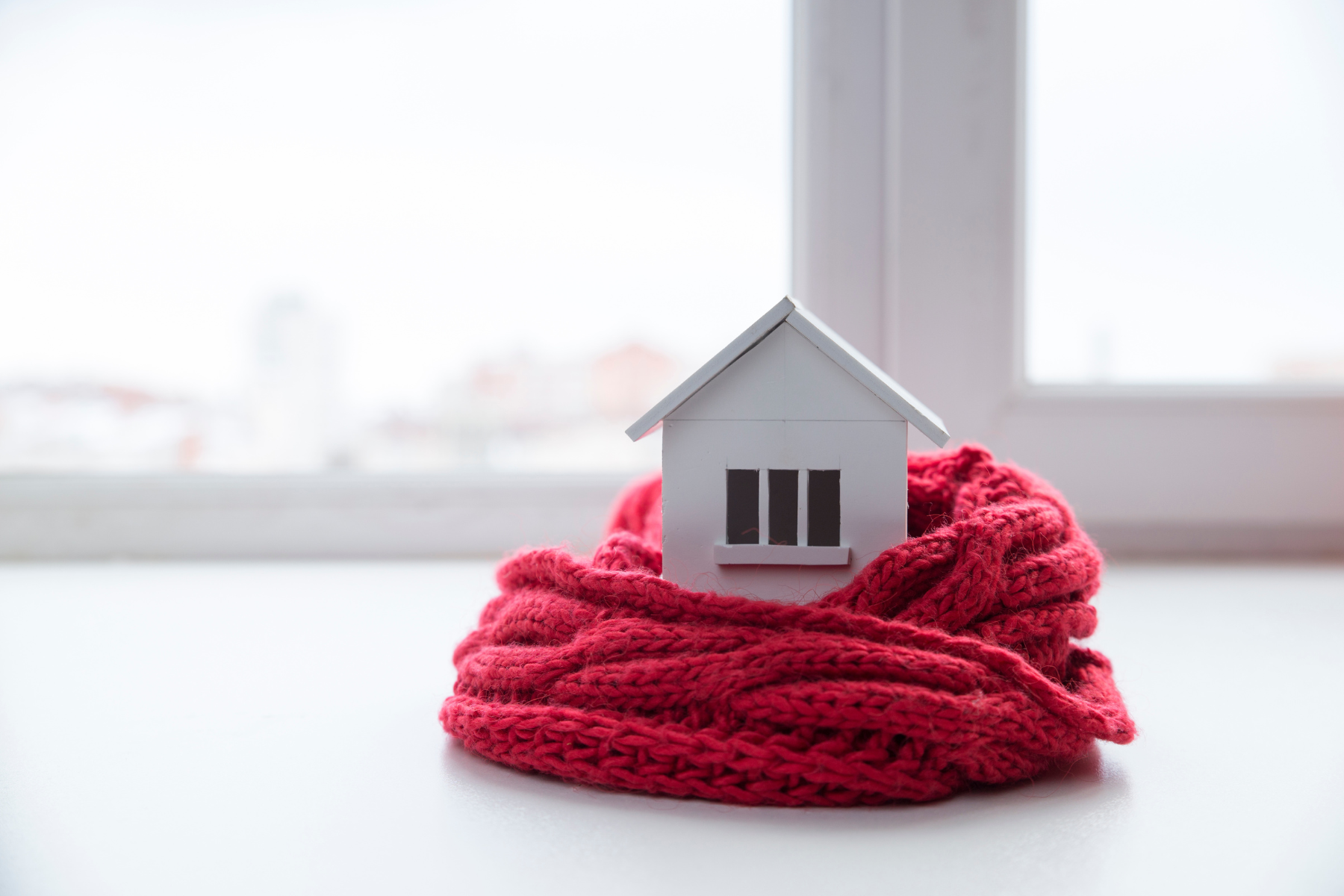A small model house wrapped in a thick red knitted scarf, representing keeping a home warm during winter.