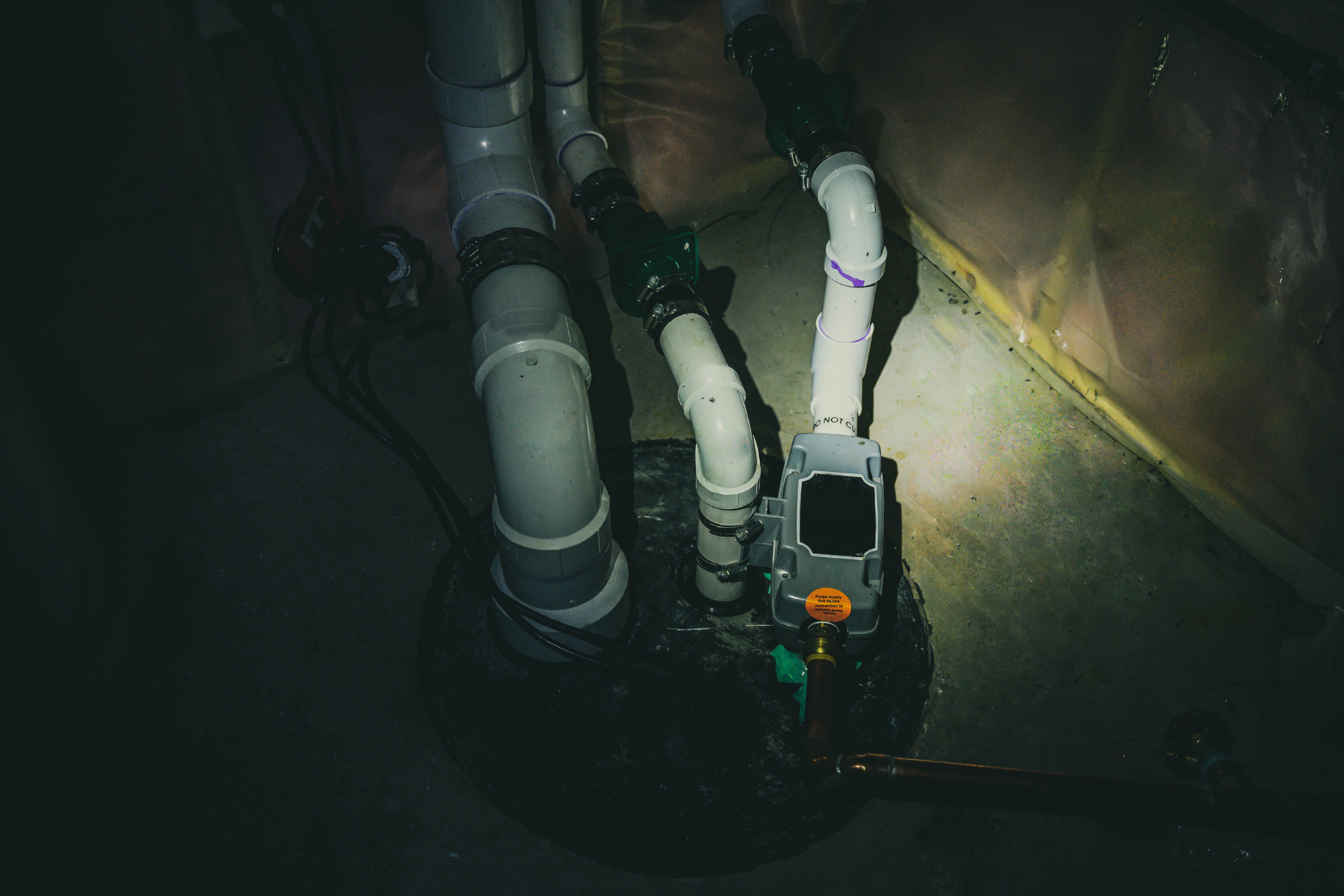 A basement sump pump and plumbing pipes installed in a dark corner.