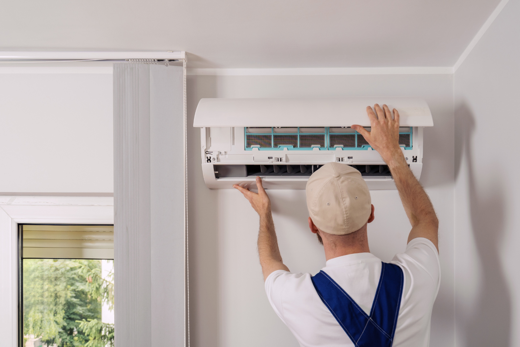 Man wearing overals and cap is checking AC unit.