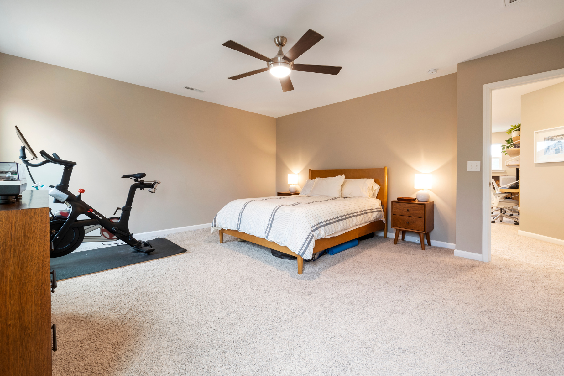 View of room with bed, bed side table, spinning bike and ceiling fan.